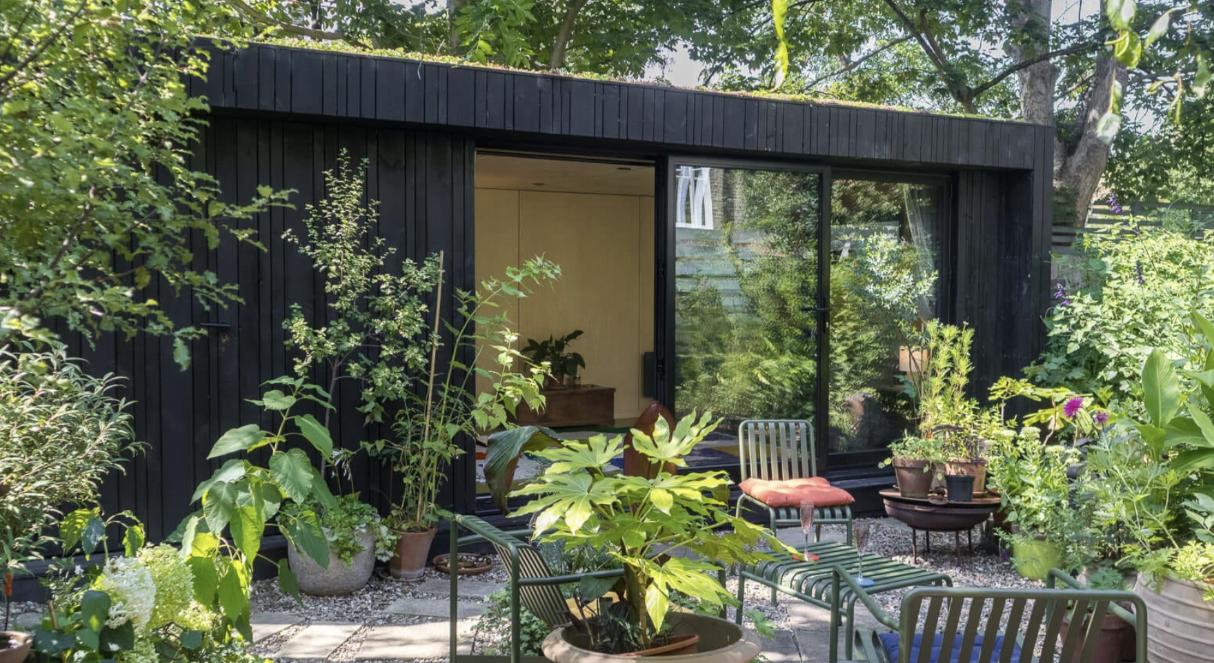 Black timber garden room with a living sedum green roof, surrounded by lush potted plants and a gravel patio with green outdoor lounge furniture.